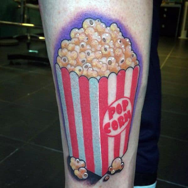 Tattoo of a striped popcorn bucket with cartoon eyes peeking from each kernel and the word POPCORN in bold red letters.