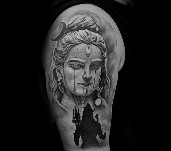 Upper arm tattoo of Lord Shiva with a calm, closed-eye expression and crescent moon, paired with a bold silhouette of Shiva holding a trident below.