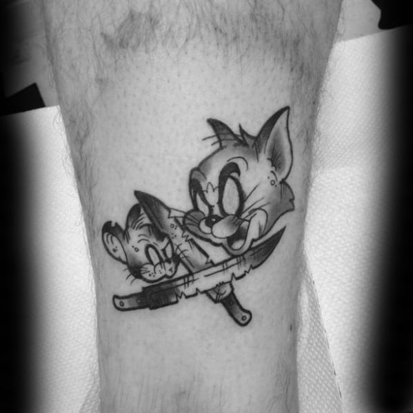 Black and grey Tom and Jerry tattoo showing Tom holding a knife with Jerry’s reflection in the blade, inked on a man’s leg.