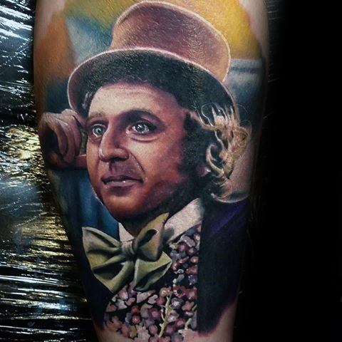 Realistic tattoo of Willy Wonka resting his head on his hand, wearing a brown top hat, green bow tie, and floral-patterned vest.