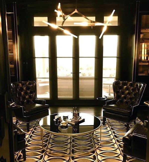 Luxurious man cave with leather chairs, patterned rug, circular glass table, and modern chandelier by large windows