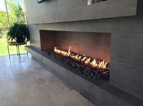 Modern outdoor fireplace with flames on pebbles, next to a potted fern on a small table, facing a grassy lawn