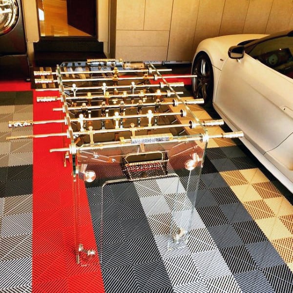 A transparent foosball table is placed in a garage next to a white car on a patterned floor
