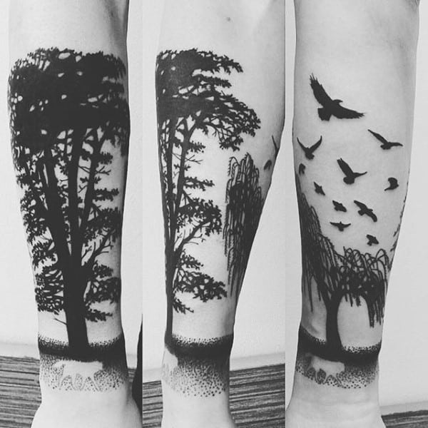 Black ink forearm tattoo featuring a forest silhouette with tall trees, a willow tree, and birds in flight, creating a serene nature scene.