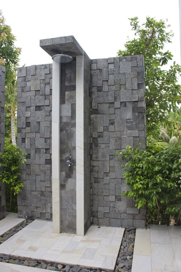 Modern outdoor shower with dark textured stone walls and white accents surrounded by greenery.