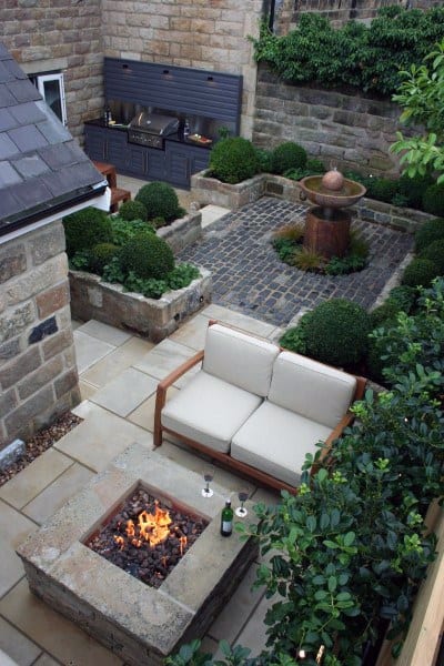 Charming stone patio with fire pit, seating, grill, and fountain nestled among lush greenery