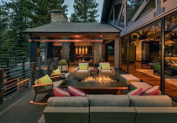 Cozy patio with a fire pit, surrounded by cushioned seating, adjacent to a glass-walled modern home in a forest setting