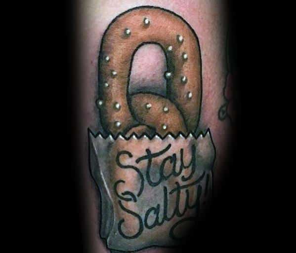 Realistic pretzel tattoo in a paper bag with "Stay Salty" written in cursive, featuring shading and salt details for depth.