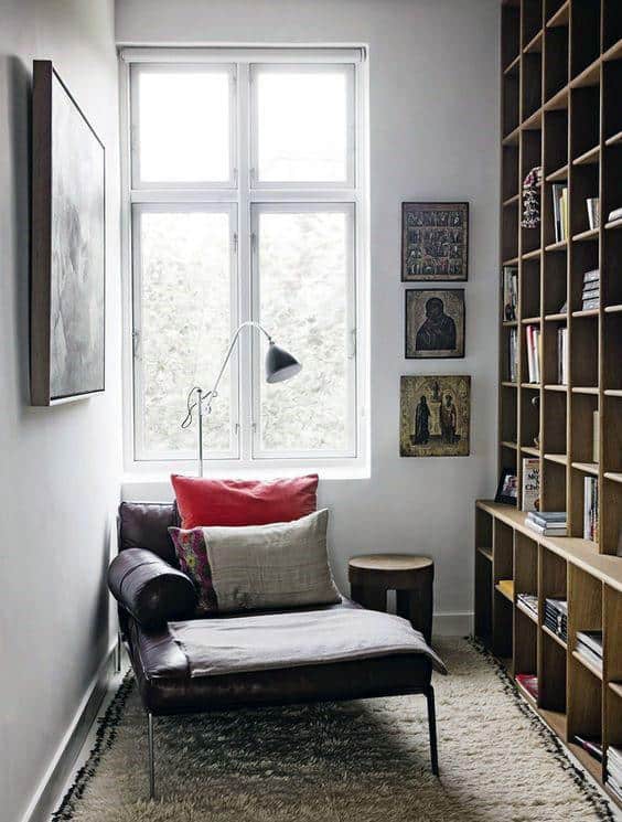 Cozy room with a chaise lounge, red and beige cushions, floor lamp, library shelves, and wall art by a window