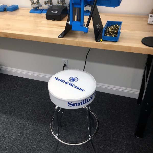 Minimalist reloading bench with a clean wooden surface, blue press setup, and a branded Smith & Wesson stool