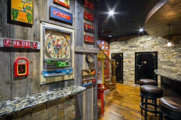 Rustic room with arcade games on wood-paneled walls, stone accents, a bar with stools, and a wooden staircase
