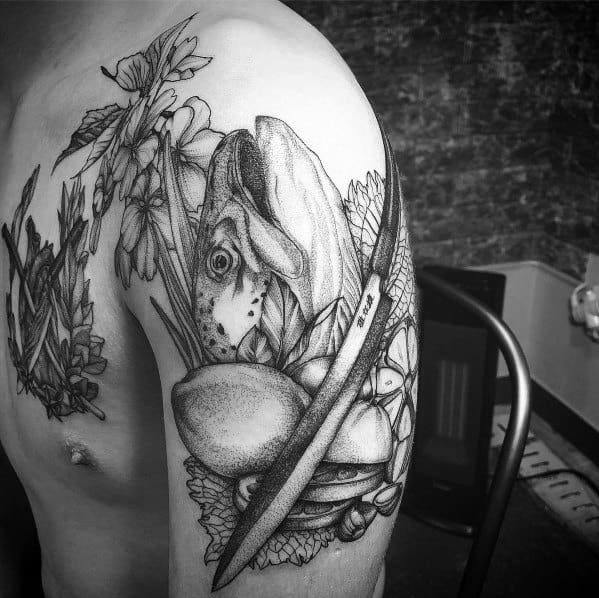 Detailed black and grey tattoo of a salmon with a fillet knife, surrounded by flowers and rocks, covering the upper arm with intricate shading.