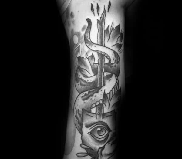 Black and grey shovel tattoo with a snake wrapped around the handle, an eye on the blade, and bold shading for a mysterious and symbolic design.