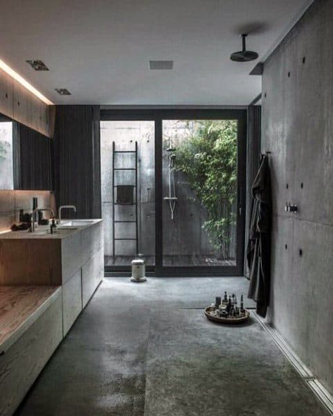 Modern bathroom with concrete walls, large glass shower door, and a leafy outdoor view. Minimalist decor and fixtures