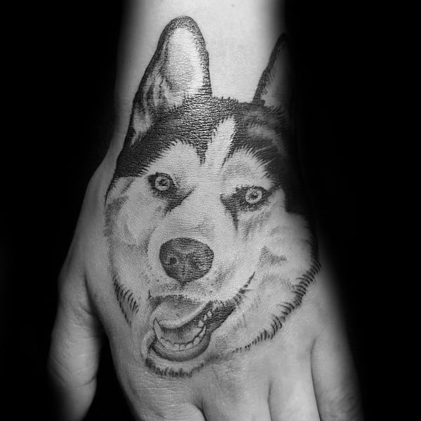 Realistic Siberian Husky tattoo on the back of a hand, featuring fine shading, expressive blue eyes, and a happy expression for a lifelike design.