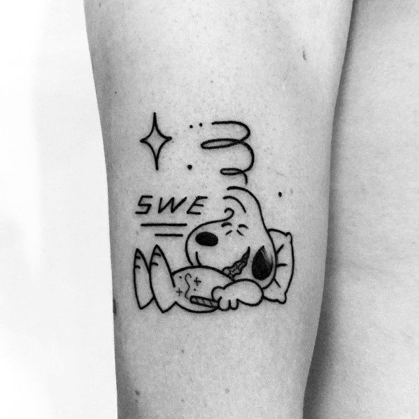 Black and white tattoo of Snoopy sleeping on a pillow with "SWE" text, motion lines, and sparkles, placed on a person's arm.