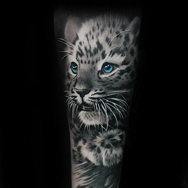 Realistic snow leopard cub tattoo in black and gray with striking blue eyes, capturing innocence and wild beauty on the forearm.