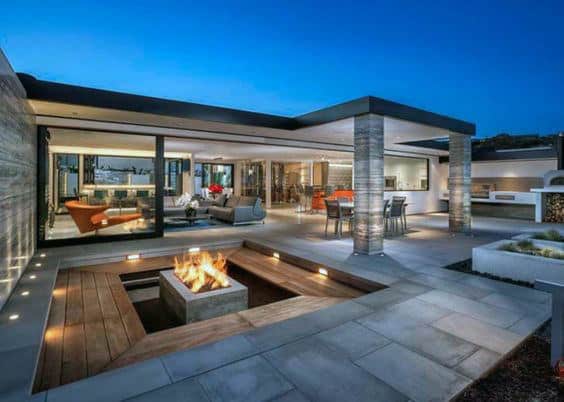 Modern patio with sunken fire pit, outdoor dining area, and glass walls connecting to a stylish interior at dusk