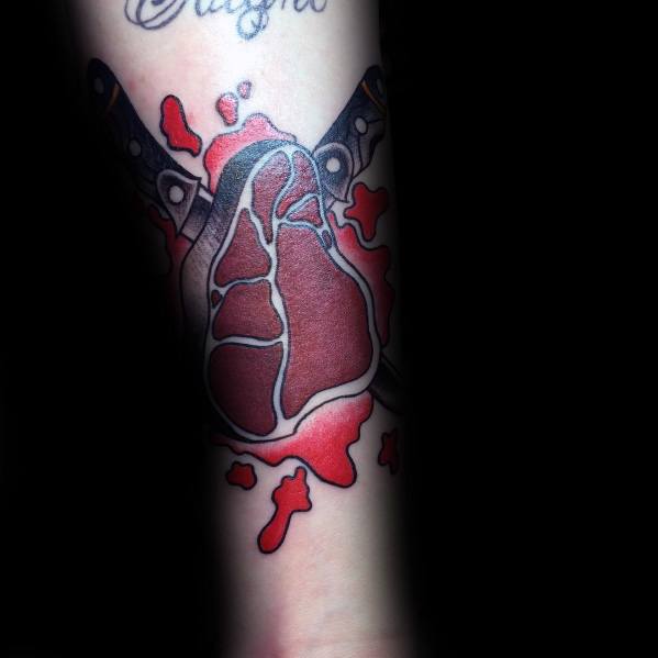 Tattoo of a steak shaped like an anatomical heart, pierced by two crossed knives, with red splatter details for a bold and dramatic effect