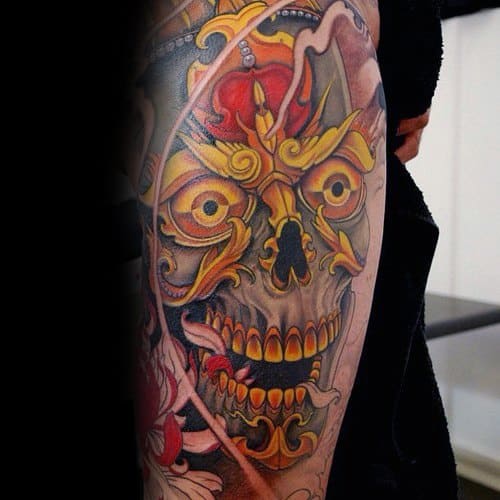 Vivid Tibetan skull tattoo on the thigh with fiery orange and yellow tones, ornate detailing, and intense facial expression.