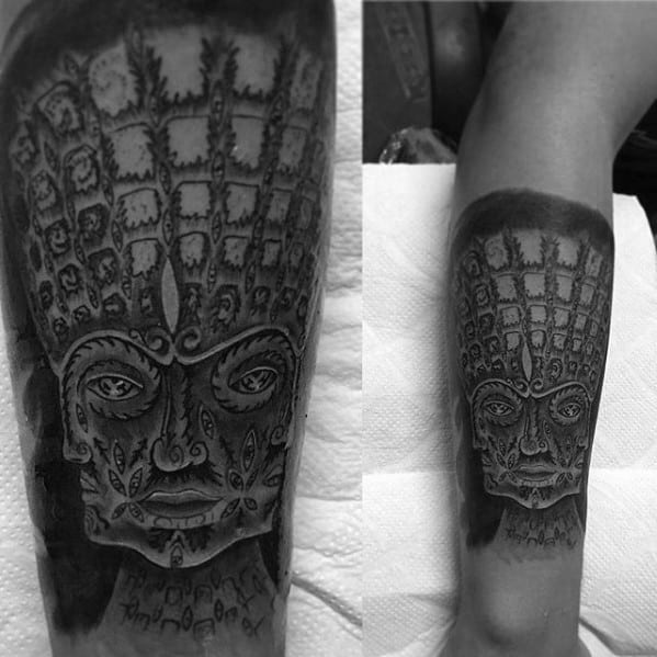 Black and grey Tool-inspired tattoo of a symmetrical face with intricate patterns and deep shadows covering the forearm.