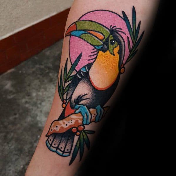 Neo-traditional toucan tattoo with bold lines, vibrant colors, and tropical leaves against a pink sun backdrop on the forearm.