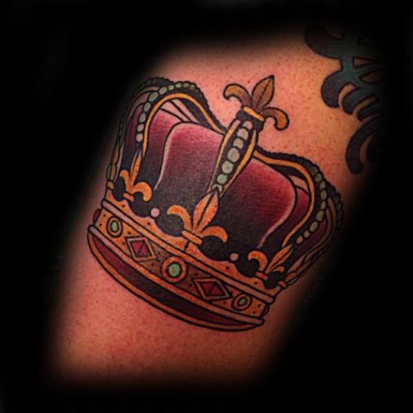 Bold traditional crown tattoo in red, gold, and green tones with jewel details, inked on the upper arm.