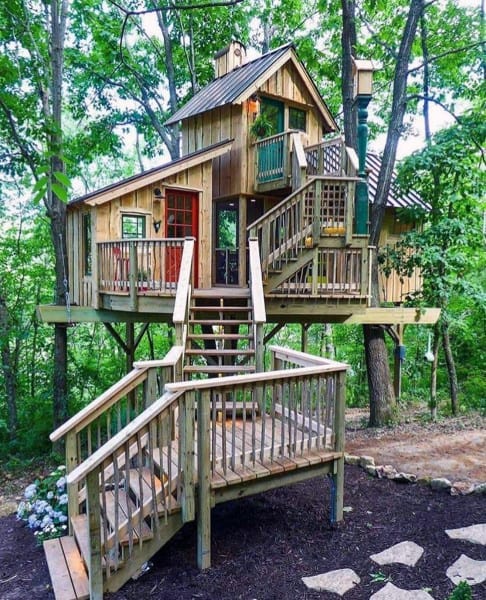 A rustic treehouse with multiple levels and wooden stairs, surrounded by lush greenery