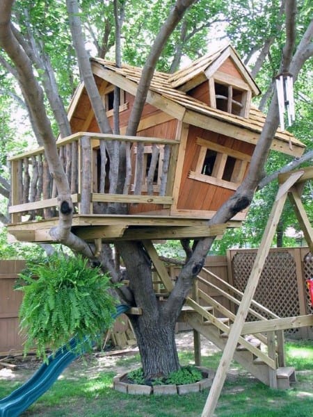 A whimsical wooden treehouse with a slanted roof, nestled among trees, with a slide and stairs