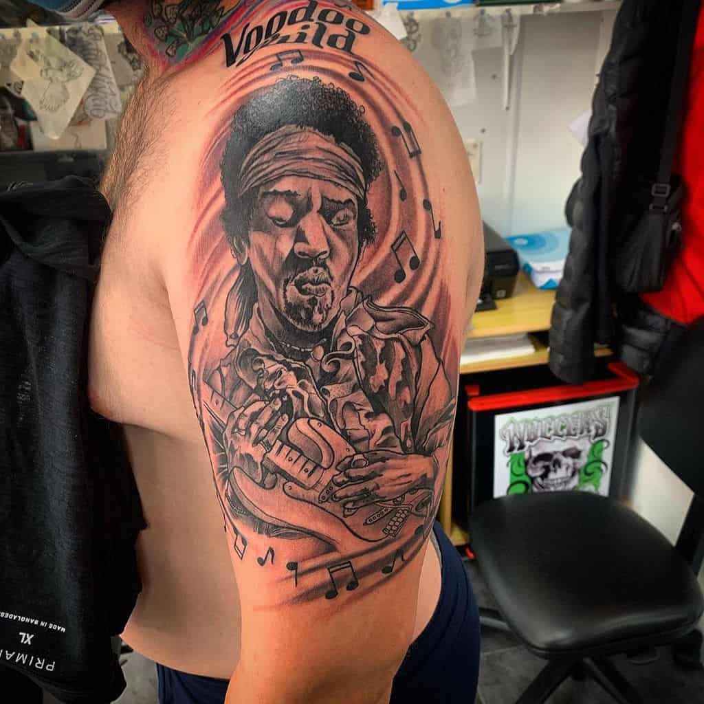 Upper arm tattoo featuring a detailed portrait of a musician in grayscale, surrounded by flowing musical notes and the text 'Voodoo' at the top