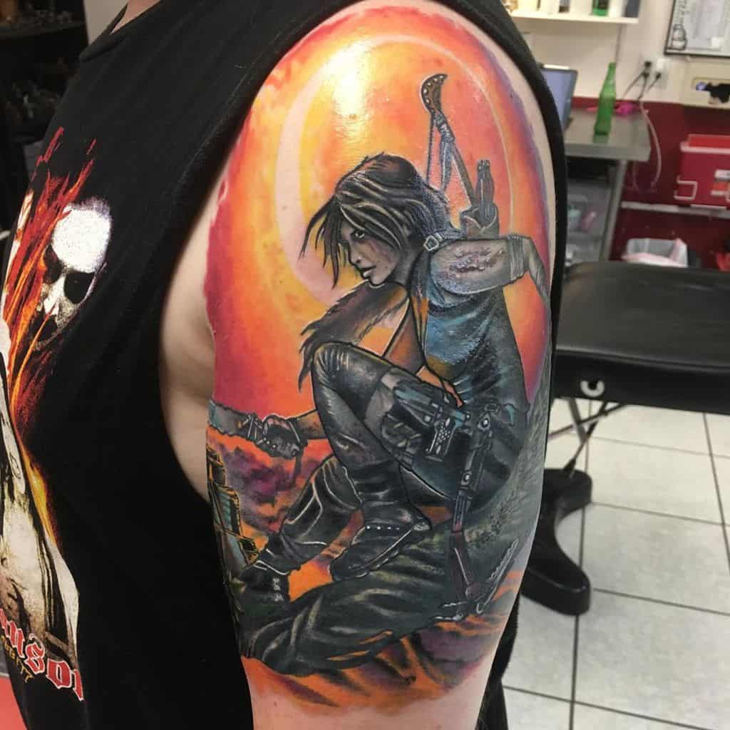 Colorful shoulder tattoo depicting a warrior woman with weapons against a vibrant orange sunset background
