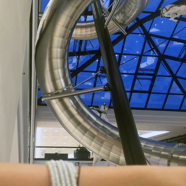 A large silver slide gracefully curves beneath a stunning glass ceiling