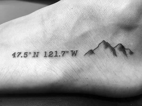 Minimalist coordinate tattoo on the foot with fine black lettering and a simple mountain design, symbolizing a meaningful outdoor location.