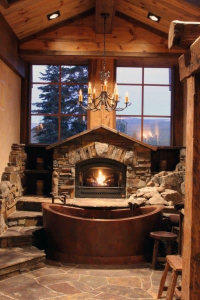 Rustic stone fireplace, bathtub, and chandelier in a cozy wooden room with a crackling fire