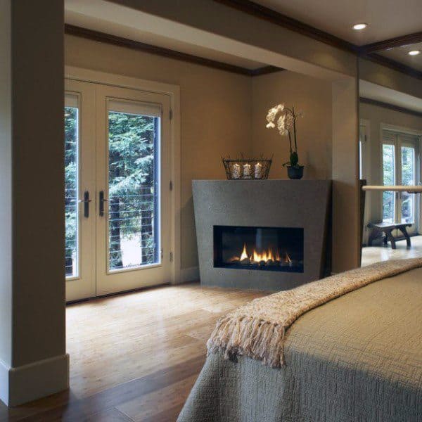 Bedroom with modern corner fireplace, cozy bed, and glass doors leading to a forest view.
