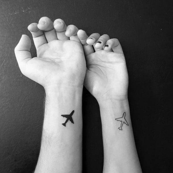 Matching wrist tattoos of airplane silhouettes, one solid and one outlined, symbolizing shared journeys, adventure, and love across distance.