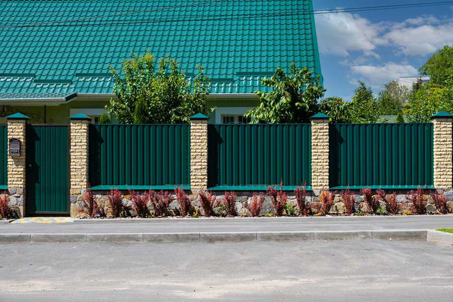 Green corrugated metal fence with brick pillars, surrounding a house with a green roof.