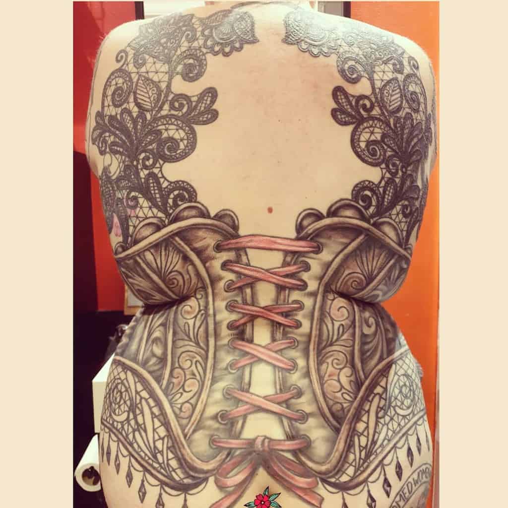 Full-back tattoo designed to resemble a detailed corset with pink ribbon lacing, ornate filigree, and intricate black lace patterns.