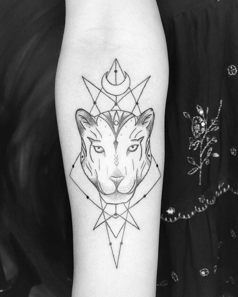 A minimalist geometric tattoo featuring the face of a jaguar, surrounded by clean lines, shapes, and celestial symbols, placed on the forearm.