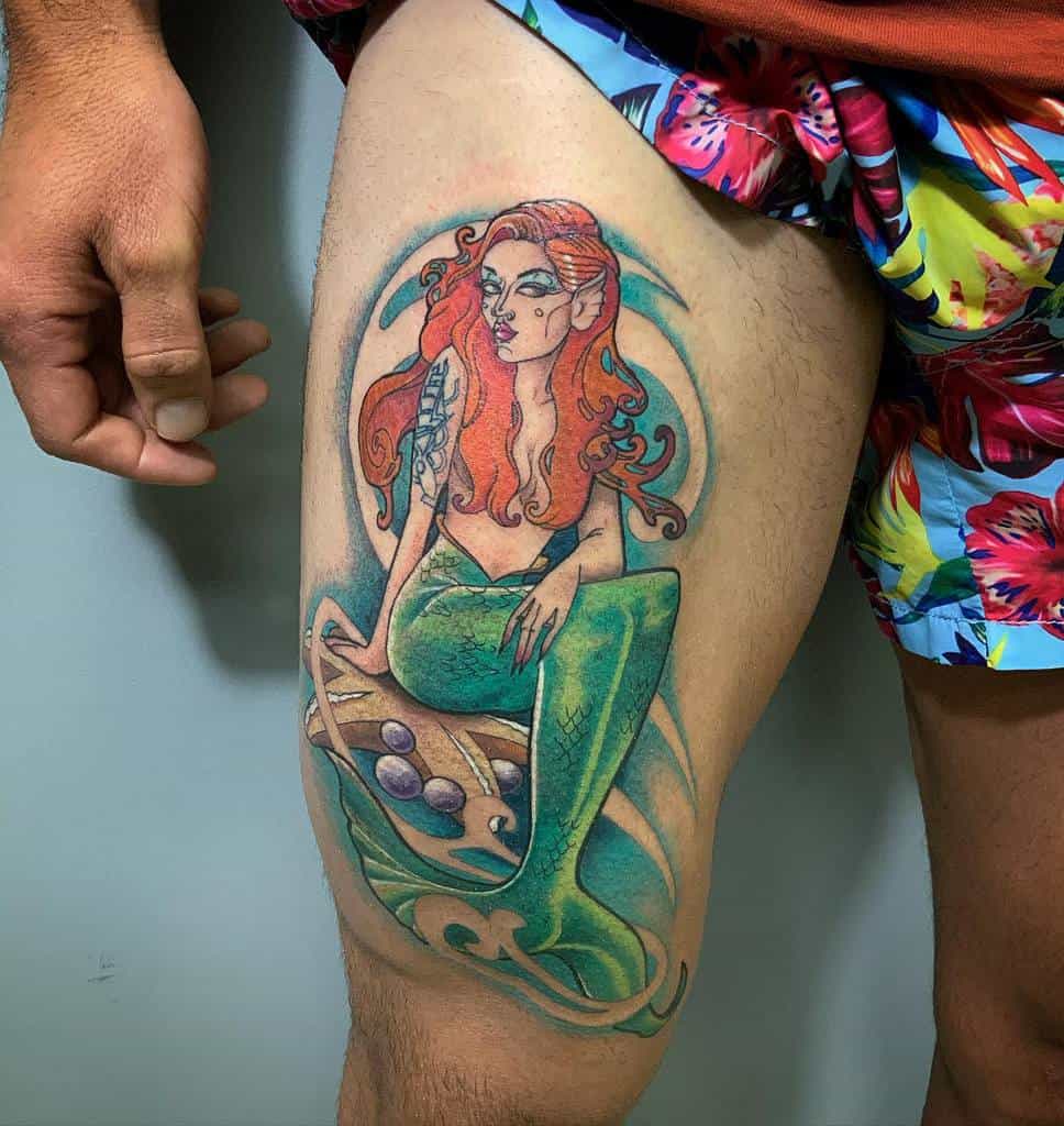 Colorful tattoo of a mermaid with vibrant red hair, green tail, and detailed fantasy elements, sitting on a rock with ocean waves around her.