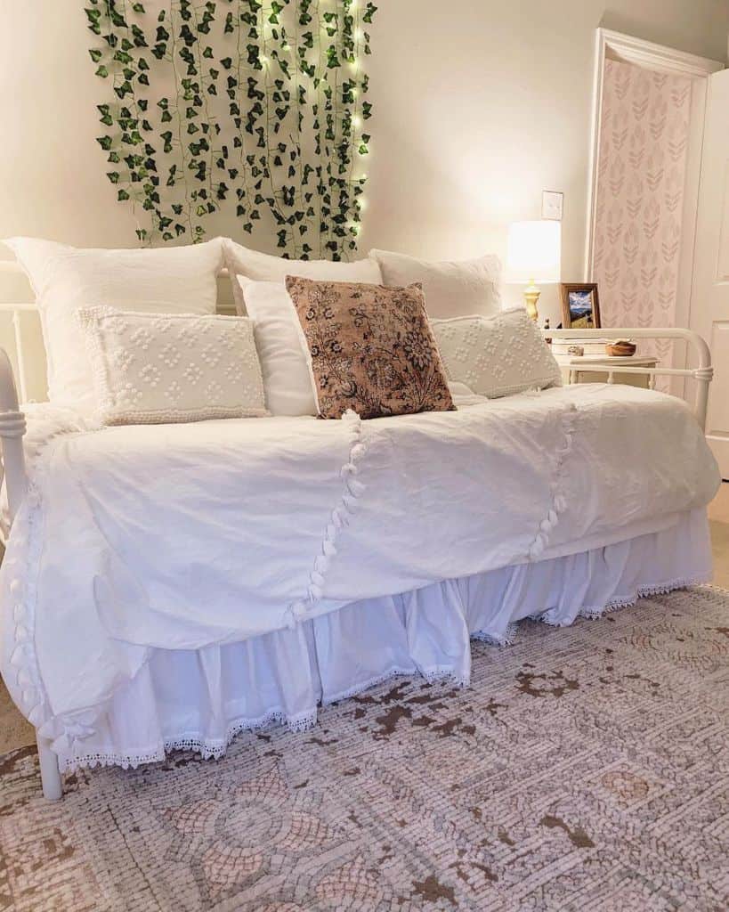 Cozy white bedroom with a daybed, patterned pillow, hanging green vines, and a small lit lamp on the bedside table