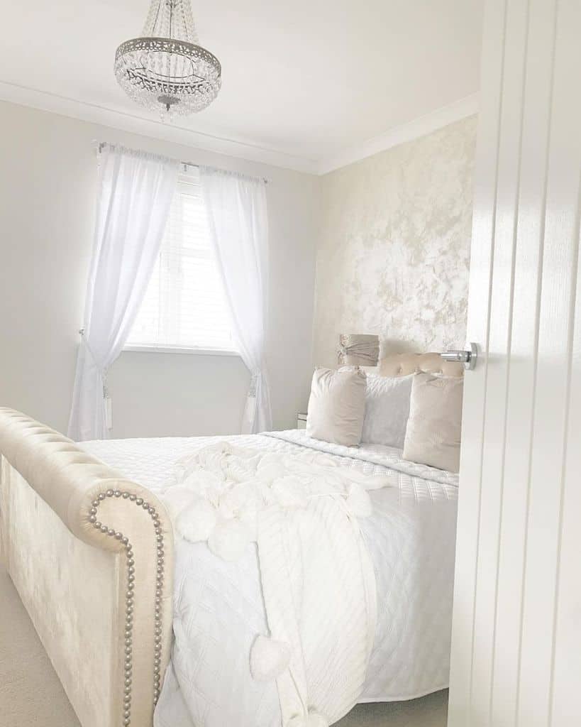 A bright bedroom featuring a white upholstered bed, crisp bedding, and sheer curtains on the window
