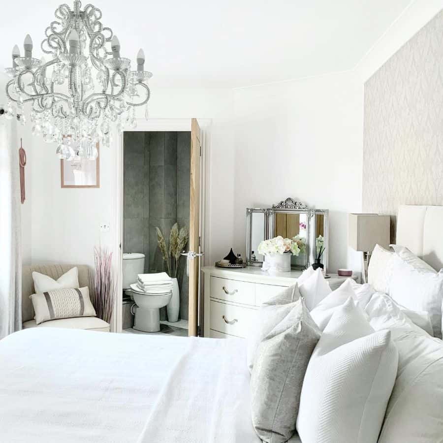 Elegant white bedroom with chandelier, pillows, and ensuite; white dresser with flowers and mirror on the side