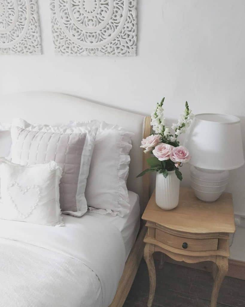 A cozy white bedroom with a pillow-laden bed, white nightstand, and a lamp beside pink and white flowers