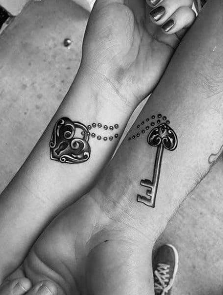 Matching wrist tattoos of a heart-shaped lock and ornate key with dotted chains, symbolizing love, trust, and a perfect match.