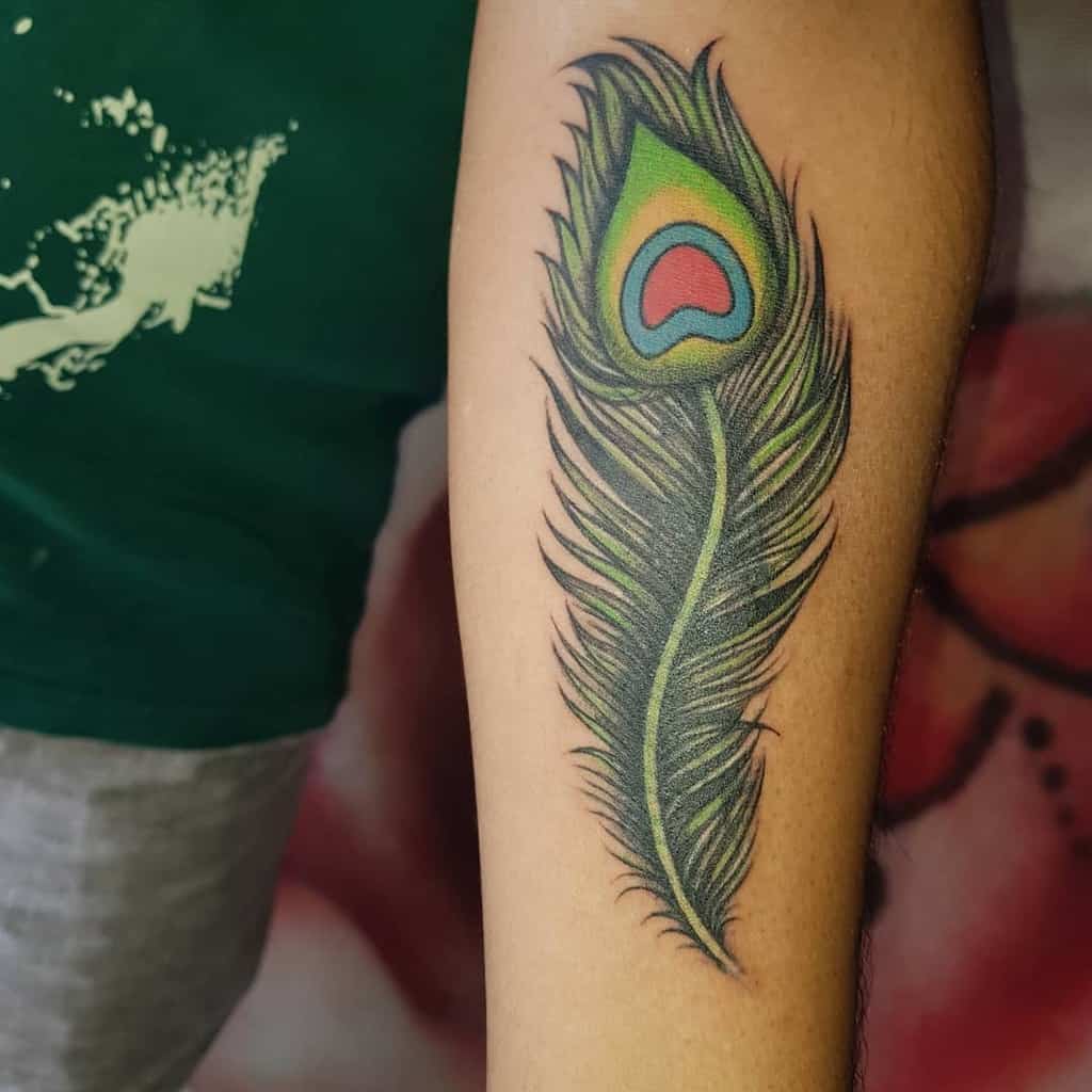 Vibrant peacock feather tattoo on the forearm, featuring bold green, blue, and red hues with detailed shading and smooth gradients.