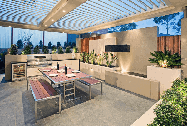 Modern outdoor kitchen with dining table, built-in grill, and plants under a pergola at dusk