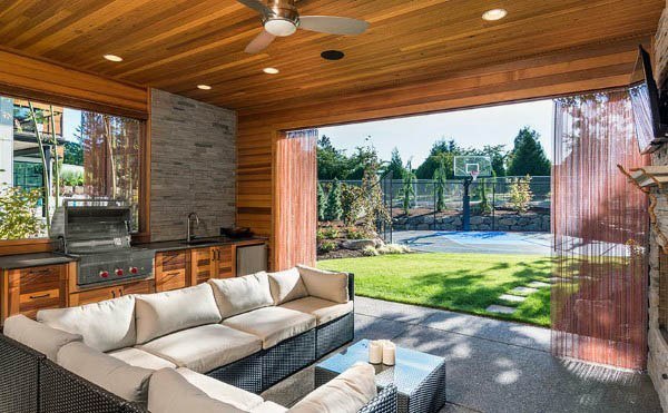 Outdoor covered patio with a sofa, grill, and ceiling fan overlooking a grassy backyard with a basketball court