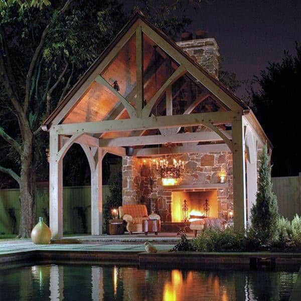 Cozy outdoor pavilion with a stone fireplace, warm lighting, and seating, next to a pool at night