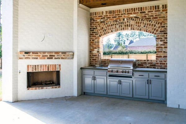 Outdoor kitchen with brick arch, built-in stainless steel grill, and gray cabinets beside a brick fireplace